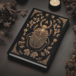 May include: A black hardcover journal with a gold beetle design. The beetle is intricately detailed with floral patterns and has a large eye in the center of its head.