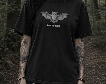 May include: Black t-shirt featuring a detailed illustration of a bat with outstretched wings. The text "I am the night" is printed below the bat. The shirt has a vintage, worn look.