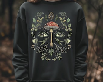 May include: Black sweatshirt featuring a nature-inspired design. The graphic includes a red-capped mushroom, eyes, and foliage arranged to resemble a face. The design incorporates leaves, branches, and a pinecone, creating a whimsical, earthy aesthetic.