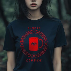 May include: A dark blue t-shirt with a red graphic design. The graphic features a coffee mug with an eye in the center, surrounded by a circle of runes and the words "SUMMON COFFEE".