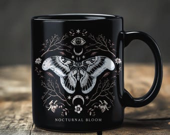May include: Black ceramic mug featuring a detailed white moth design with celestial elements. The design includes an eye, crescent moons, stars, and floral accents. The words "NOCTURNAL BLOOM" are printed at the bottom of the mug.