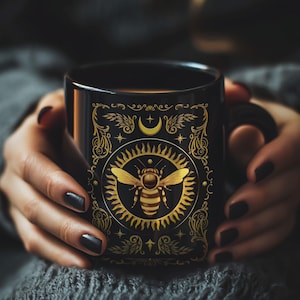 May include: Black ceramic mug with a gold design featuring a bee, sun, moon, and stars. The mug is being held by a person's hands.