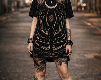 May include: Black t-shirt dress with a detailed gold antler and bone design. The dress features a crescent moon and star graphic at the neckline. The model is wearing black boots and bracelets. The background is a blurred city street.