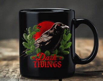 May include: Black ceramic mug featuring a detailed illustration of a raven with red eyes, set against a red sun and green foliage. The mug has the words "Dark Tidings" in red script.