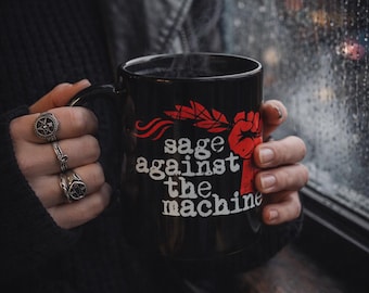 May include: Black coffee mug with the text "sage against the machine" in white and red. The mug has a red fist graphic and a feather design. The mug is held by a person wearing rings and a black sweater.