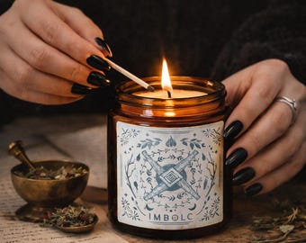 May include: A lit amber glass candle with a white label featuring a blue and white design and the word "IMBOLC". A match is being used to light the candle. A small brass bowl and spoon are visible.