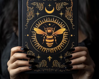 May include: A black journal with a gold bee design on the cover. The bee is surrounded by a sun and moon design with stars and floral accents.