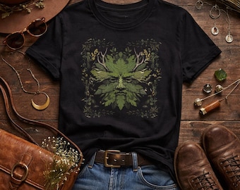 May include: Black t-shirt featuring a green leaf-based design of a face with antlers. The design is surrounded by leaves and foliage. The shirt is paired with blue jeans, a brown belt, and brown leather boots. A brown leather purse and accessories are also in the image.