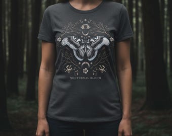 May include: Dark gray t-shirt featuring a detailed graphic of a moth with intricate wing patterns, surrounded by celestial and floral elements. The text "NOCTURNAL BLOOM" is printed below the design. The shirt is made of a soft material.