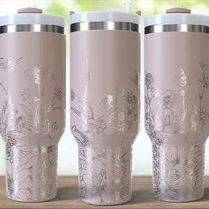 May include: Three insulated tumblers with handles, featuring a light pink finish and silver floral designs. Each tumbler has a white lid and a matching pink handle. The tumblers are displayed on a wooden surface.