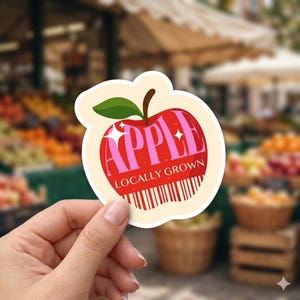 May include: A sticker in the shape of a red apple with the word "APPLE" in pink letters. The words "LOCALLY GROWN" are below the apple. The sticker has a cream-colored border and a green leaf.