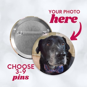 May include: Round button pin with a photo of a black dog wearing a blue and red plaid bow tie. The text "YOUR PHOTO here" and "CHOOSE 3-9 pins" are in pink. The pin has a metal back and a safety pin clasp.