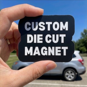 May include: A black custom die-cut magnet with white text is held up against a blue sky. The magnet's rounded rectangular shape is visible, with the words "CUSTOM DIE CUT MAGNET" in bold, white letters. A car is visible in the background.
