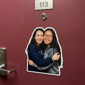 May include: A sticker of two smiling women hugging, affixed to a maroon door. One woman wears a navy blue sweatshirt, the other a gray hoodie and glasses. Above, a silver plate reads "113".