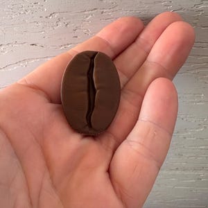 May include: A dark brown, oval-shaped coffee bean. The bean has a deep groove down the center, and is held in a person's hand. The bean is a solid color with a smooth texture.
