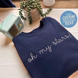 May include: Navy blue sweatshirt with white embroidered text "oh my stars!". A light blue retro radio and a small potted plant are in the background. A blue circle with the text "YOUR CUSTOM EMBROIDERED TEXT" is also visible.