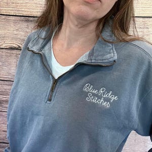 May include: A dusty blue pullover sweatshirt with a quarter-zip neckline and the embroidered text "Blue Ridge Stitches" in white script. The sweatshirt has a relaxed fit and a white undershirt is visible underneath.