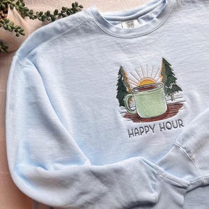 Peut inclure: Un sweat-shirt bleu clair avec un motif brodé représentant une tasse verte, des arbres et un soleil levant. Les mots "HAPPY HOUR" sont brodés en dessous. Le sweat-shirt est en matière douce.