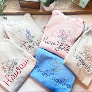 May include: A collection of embroidered sweatshirts in various colors, each featuring a state outline and floral or botanical designs. States include Texas, New York, Hawaii, North Carolina, and Virginia. The text of each state is embroidered in a complementary color.