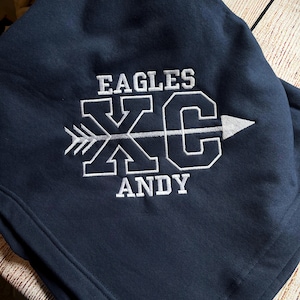 May include: A navy blue hooded sweatshirt featuring white embroidery. The design includes the text EAGLES, the letters XC with an arrow running horizontally through them, and the name ANDY embroidered in white thread below the graphic.