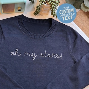May include: A dark navy blue long-sleeve sweater with a ribbed crew neckline. The front of the sweater features white embroidered cursive text that reads "oh my stars!" in a simple, clean font style across the chest area of the garment.