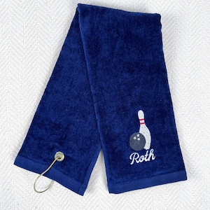May include: A navy blue bowling towel with a white and red bowling pin and a black bowling ball embroidered on it. The name "Roth" is embroidered in white script below the bowling ball. A metal loop is attached to one end.