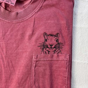 A red t-shirt with a black embroidered image of a squirrel on the front pocket.