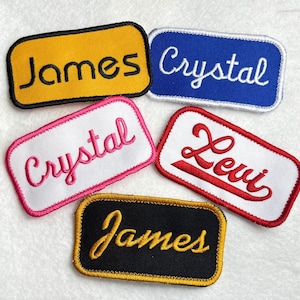 May include: Five rectangular embroidered patches with rounded corners. One yellow patch with black text James, one blue patch with white text Crystal, one white patch with pink text Crystal, one white patch with red text Levi, and one black patch with yellow text James.