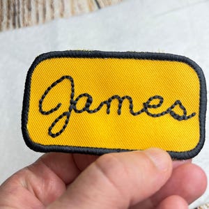 May include: A rectangular embroidered patch with a bright yellow background and a thick black border. The name James is stitched in the center in a black cursive font. The patch has rounded corners and a visible fabric texture throughout the surface.