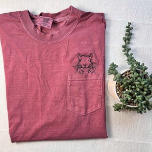 a red t-shirt with a small black squirrel embroidered on the front pocket, placed next to a potted plant on a white surface.