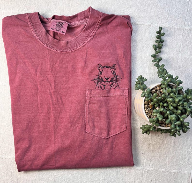 a red t-shirt with a small black squirrel embroidered on the front pocket, placed next to a potted plant on a white surface.