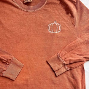 A long-sleeved orange t-shirt with a white pumpkin embroidered on the front.