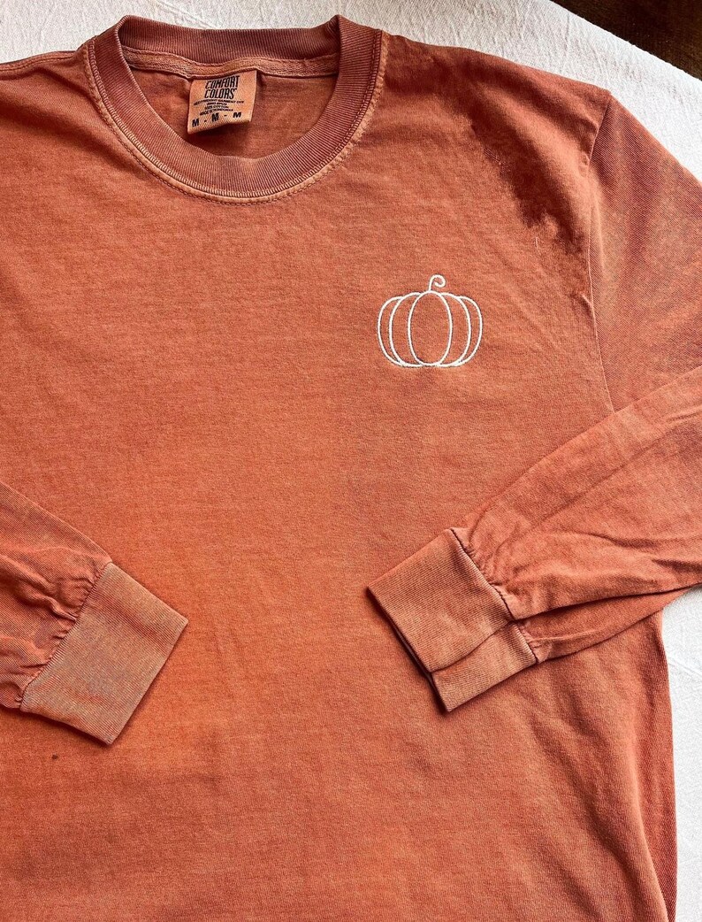 A long-sleeved orange t-shirt with a white pumpkin embroidered on the front.