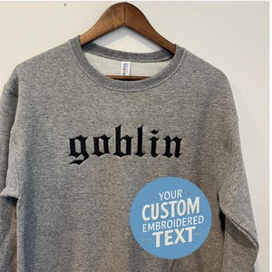 May include: A heather gray sweatshirt with the word "goblin" embroidered in black gothic lettering. A blue circle with the text "YOUR CUSTOM EMBROIDERED TEXT" is on the lower right. The sweatshirt is hanging on a wooden hanger.