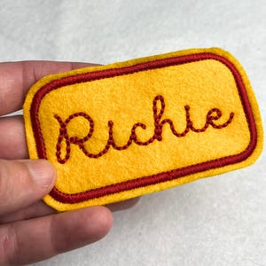 May include: A rectangular yellow felt patch with a dark red embroidered border and the name Richie stitched in dark red cursive script across the center. The patch features rounded corners and a textured fabric surface.