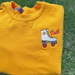 May include: Mustard yellow sweatshirt with a roller skate patch. The patch features a white skate with red laces and wheels, a gold star, and the name "Roth" in red script.