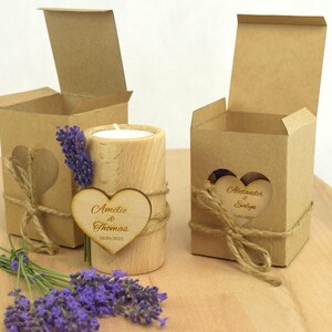 May include: Two small, brown cardboard boxes with heart-shaped cutouts and a wooden candle holder with a heart-shaped wooden tag. The tag says "Amelie & Thomas 18.09.2022". The boxes are tied with twine and have a small sprig of lavender flowers.