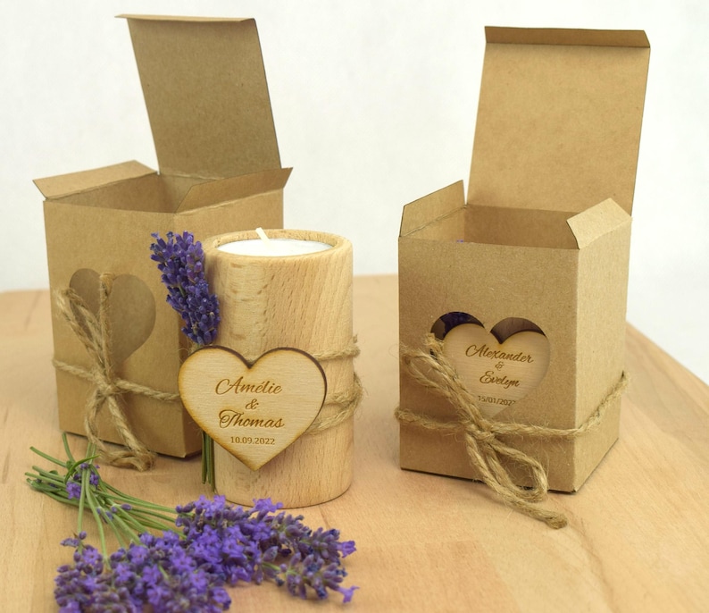 May include: Two small, brown cardboard boxes with heart-shaped cutouts and a wooden candle holder with a heart-shaped wooden tag. The tag says "Amelie & Thomas 18.09.2022". The boxes are tied with twine and have a small sprig of lavender flowers.