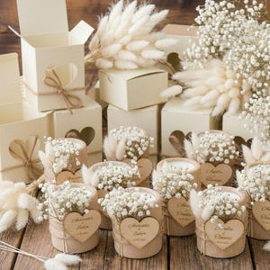 Könnte beinhalten: Eine Ansammlung cremefarbener Geschenkboxen und kleiner Holzkerzenhalter. Die Boxen haben herzförmige Ausschnitte und sind mit Bindfäden gebunden. Die Kerzenhalter sind mit weißen Blumen und Holzherzanhängern mit Namen und Daten verziert. Trockenblumen sind ebenfalls vorhanden.