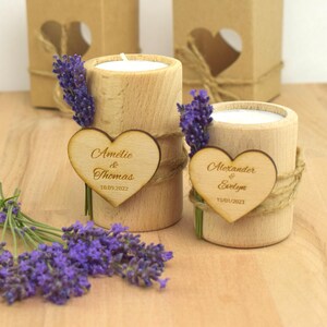 May include: Two wooden candle holders with heart-shaped wooden tags. The tags are engraved with the names "Amelie & Thomas" and "Alexander & Evelyn" along with their wedding dates. The candle holders are decorated with lavender sprigs and twine.