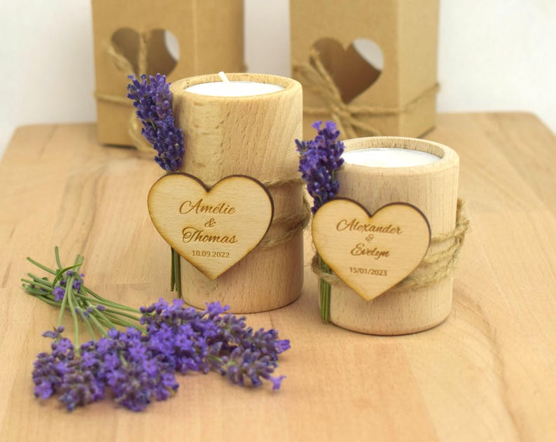 May include: Two wooden candle holders with heart-shaped wooden tags. The tags are engraved with the names "Amelie & Thomas" and "Alexander & Evelyn" along with their wedding dates. The candle holders are decorated with lavender sprigs and twine.