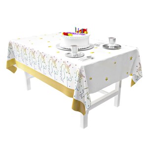 May include: White plastic tablecloth with a gold border and a unicorn design. The tablecloth has a rainbow color scheme and features small gold stars. A white cake with pink and red frosting and candles is on the tablecloth.