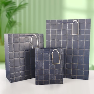 May include: Three gift bags in varying sizes. The bags are dark blue with a grid pattern and gold accents. Each bag has a gold tag and a dark blue rope handle. The bags are on a white surface.