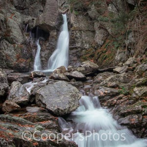 Puede incluir: Una cascada cae sobre rocas cubiertas de musgo en un entorno boscoso exuberante. El agua es blanca y espumosa, creando una sensación de movimiento y energía. Las rocas son grises y erosionadas, añadiendo una sensación de antigüedad e historia a la escena.