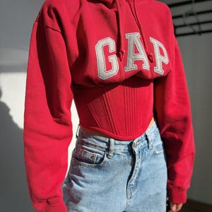 May include: A bright red cropped hooded sweatshirt featuring the word GAP in large grey block letters across the chest. The garment has long sleeves, a drawstring hood, and a structured corset-style bodice with vertical stitching details.