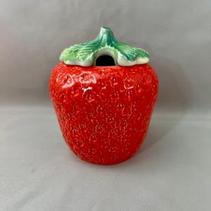 May include: A ceramic strawberry-shaped jar with a green leaf-shaped lid. The jar is a vibrant red color with a textured surface. The lid has a small opening and is painted with green and white details. This decorative jar is perfect for kitchen decor.