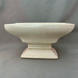 May include: A cream-colored ceramic pedestal bowl featuring a wide, flared rectangular opening that tapers down to a stepped, square base. The surface displays a visible crackle glaze finish throughout the entire exterior of the vessel.