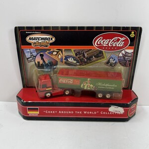 Pode incluir: Caminhão de brinquedo da marca Matchbox Collectibles Coca-Cola. A cabine vermelha está conectada a um trailer vermelho e verde com as palavras "Herbstfreuden warten auf Dich!". A embalagem diz "Coke Around the World" e apresenta a bandeira alemã.