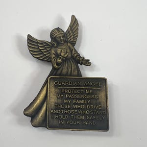Könnte beinhalten: Eine bronzefarbene Schutzengel-Figur mit ausgebreiteten Flügeln und einem rechteckigen Schild. Auf dem Schild steht "GUARDIAN ANGEL PROTECT ME MY PASSENGERS. MY FAMILY THOSE WHO DRIVE AND THOSE WHO STAND - HOLD THEM SAFELY IN YOUR HAND."