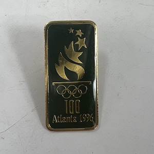 May include: A rectangular pin badge with a black background and gold trim. It features the Olympic rings, a torch design, two stars, and the text "100 Atlanta 1996" in gold. A commemorative item.